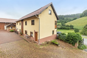 Ein zweigeschossiges Gebäude mit geneigtem Dach, einer gepflasterten Auffahrt und Blick auf einen grünen Hügel.