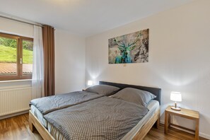 Ein Schlafzimmer mit einem Bett, einem Nachttisch, einer Lampe, einem Fenster mit Vorhängen und einem Bild an der Wand.