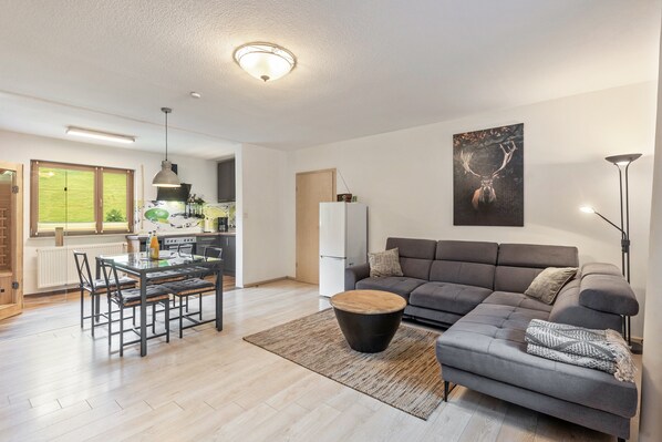 Ein modernes Wohnzimmer mit einem grauen Sofa, einem Esstisch mit Stühlen, einer Küche im Hintergrund und einem großen Hirschbild an der Wand.