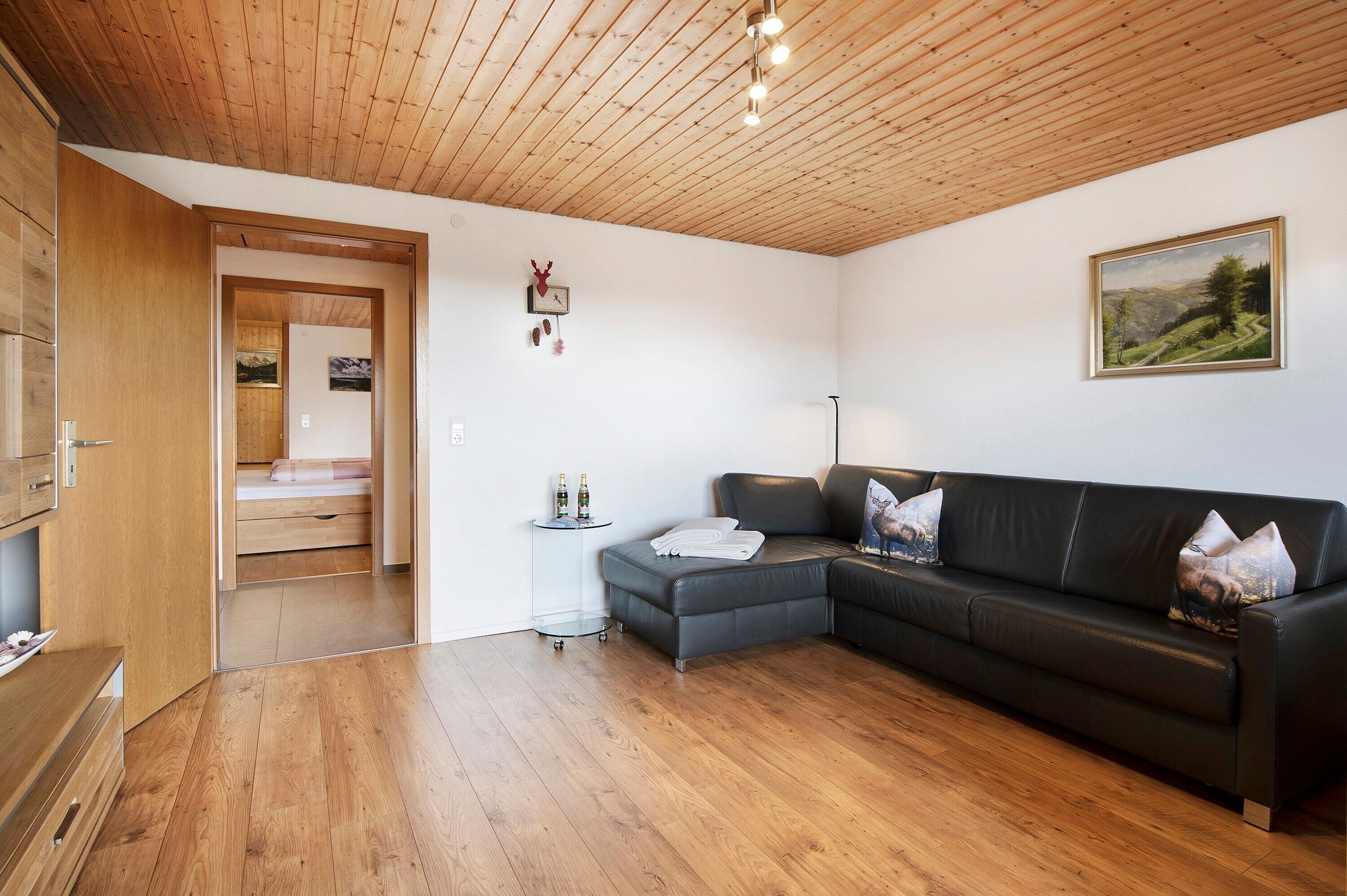 Een moderne woonkamer met een houten plafond, een leren bank in het zwart, een glazen salontafel en een schilderij aan de wand.