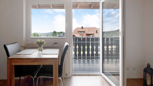 Een kamer met een houten tafel, stoelen, een vaas met bloemen en een balkon met een metalen hekwerk.
