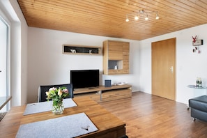 A modern living room with a wooden ceiling, a flat-screen TV, a dining table with a vase of flowers, and a built-in wooden cabinet.