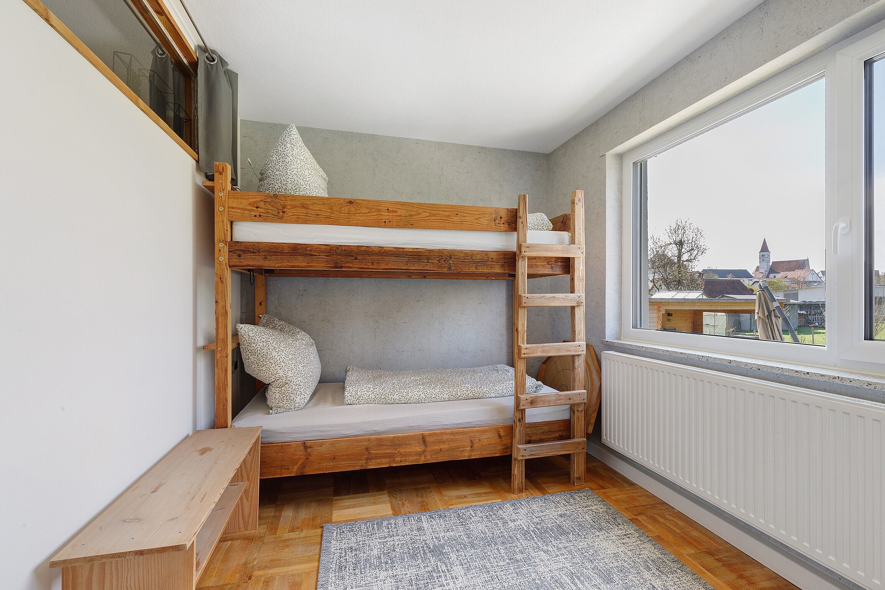 A bunk bed with a ladder, a wooden bench, a radiator, and a view through a window.