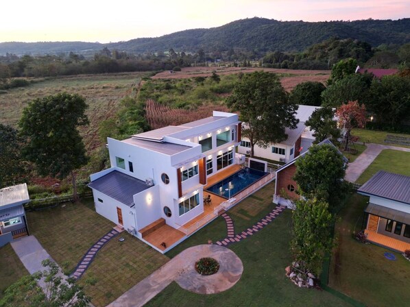 Una casa moderna de dos pisos con una amplia piscina, rodeada de césped cuidado y árboles.