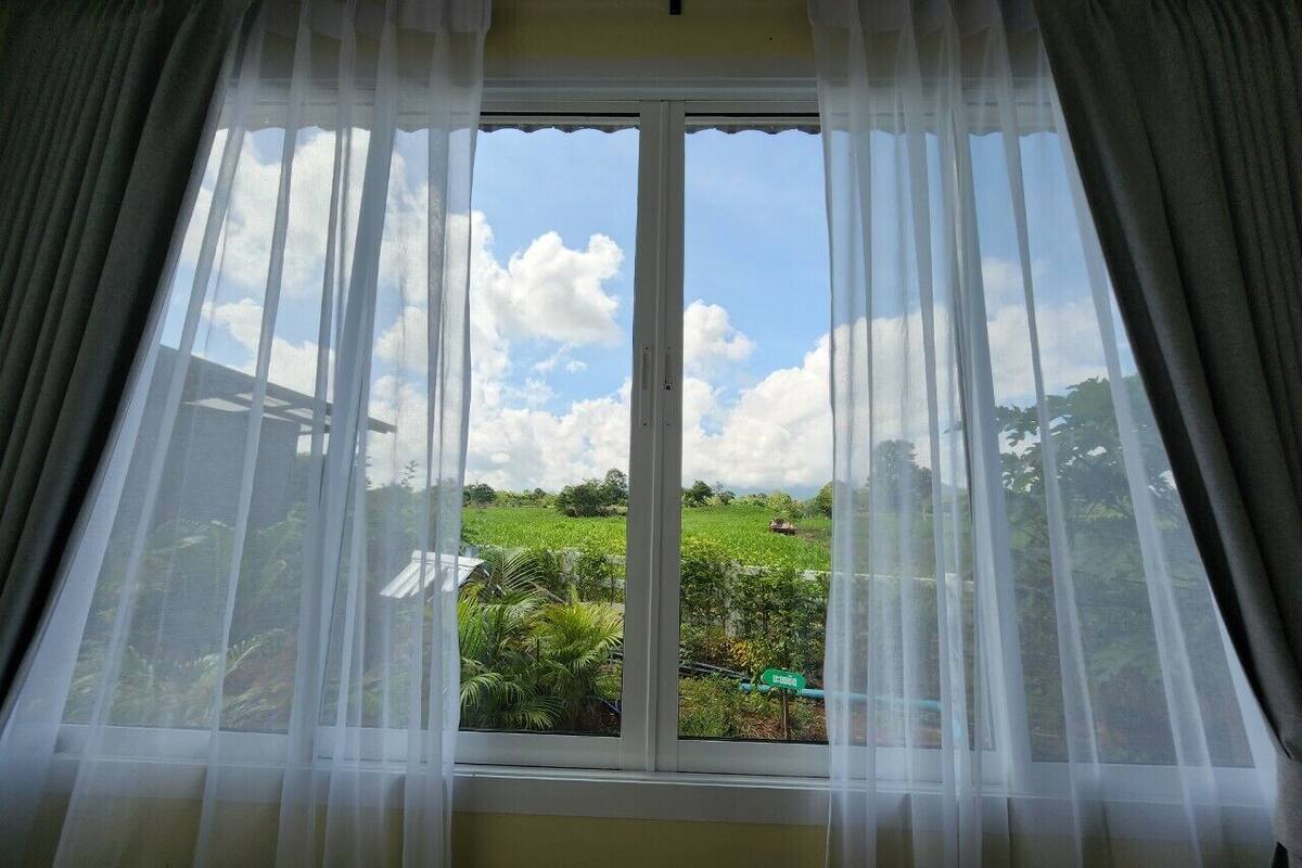 A room with a large window featuring sheer white curtains, a view of a green field, and a clear sky.
