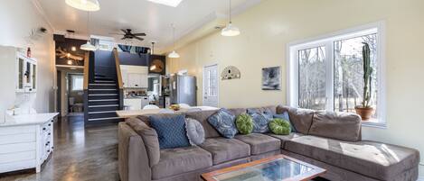 A spacious living area with a large sectional sofa, a glass coffee table, and pendant lighting. A kitchen is visible in the background with modern appliances and a staircase leading to an upper level.