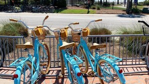Trois vélos bleus avec des selles et des paniers marron, garés sur une surface en briques près d’une clôture.
