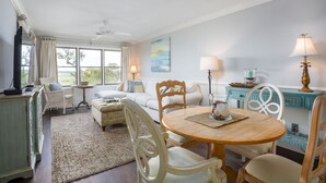 Un coin repas avec une table ronde en bois, des chaises à revêtement blanc et une vue sur l’extérieur grâce à de grandes fenêtres.