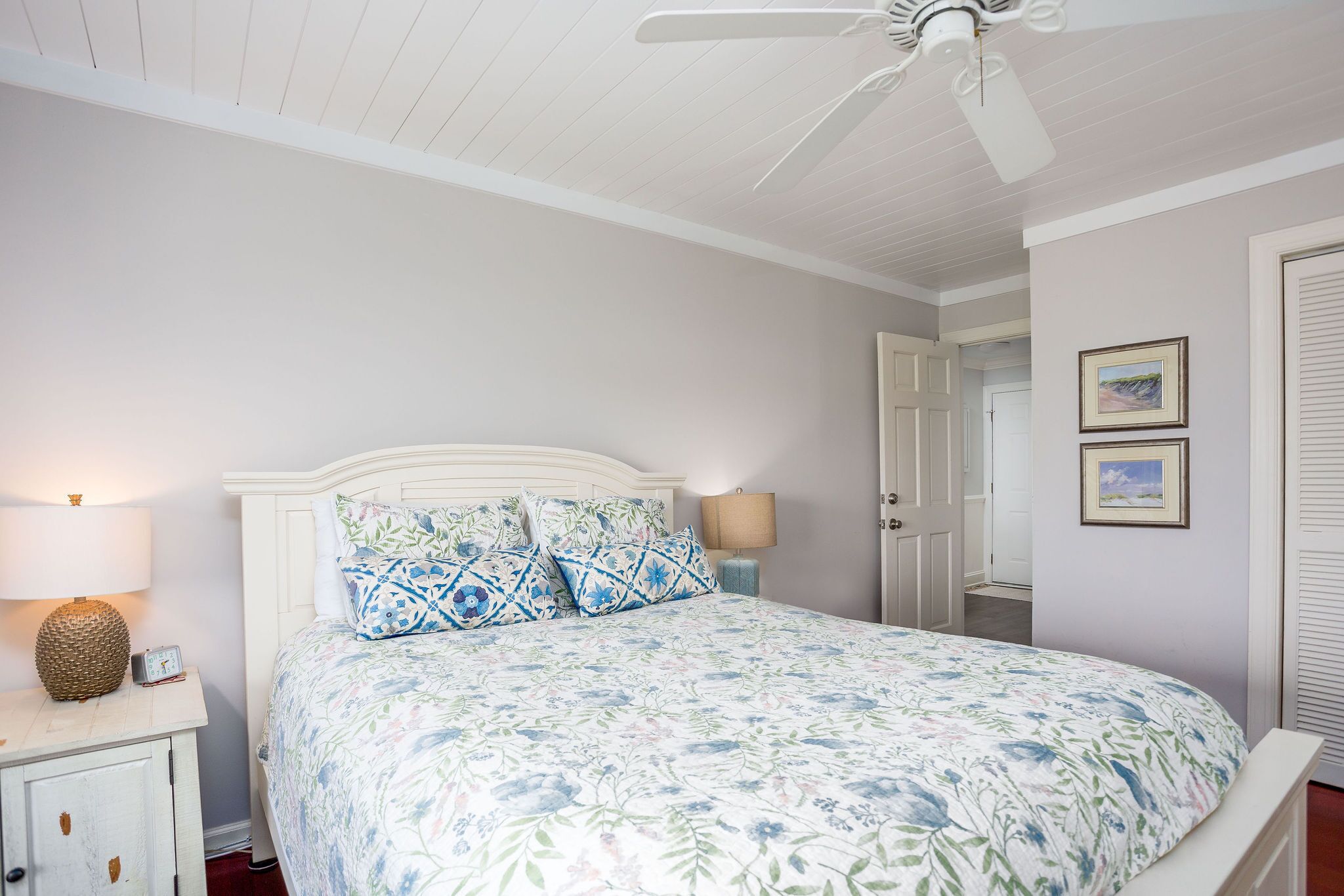 A bedroom with a bed, bedside lamps, a ceiling fan, and a door leading to another room.