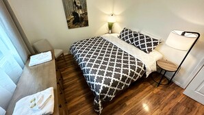 Un dormitori amb un llit, una tauleta de nit, una làmpada, una cadira i un quadre a la paret.