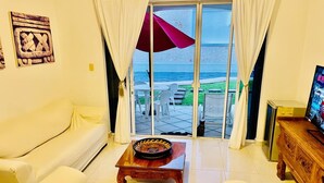 Sala de estar con un sofá blanco, una mesa de centro de madera y un televisor sobre un mueble de madera. Hay una puerta corrediza de vidrio que da a un área exterior con piscina y sillas.