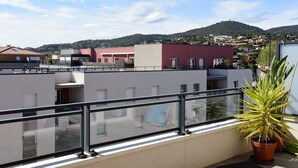 A rooftop terrace with a glass balustrade, potted plants, and a view of surrounding buildings and hills.
