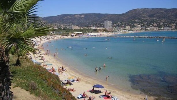 A beach with clear blue water, people sunbathing, and a mountain in the background.