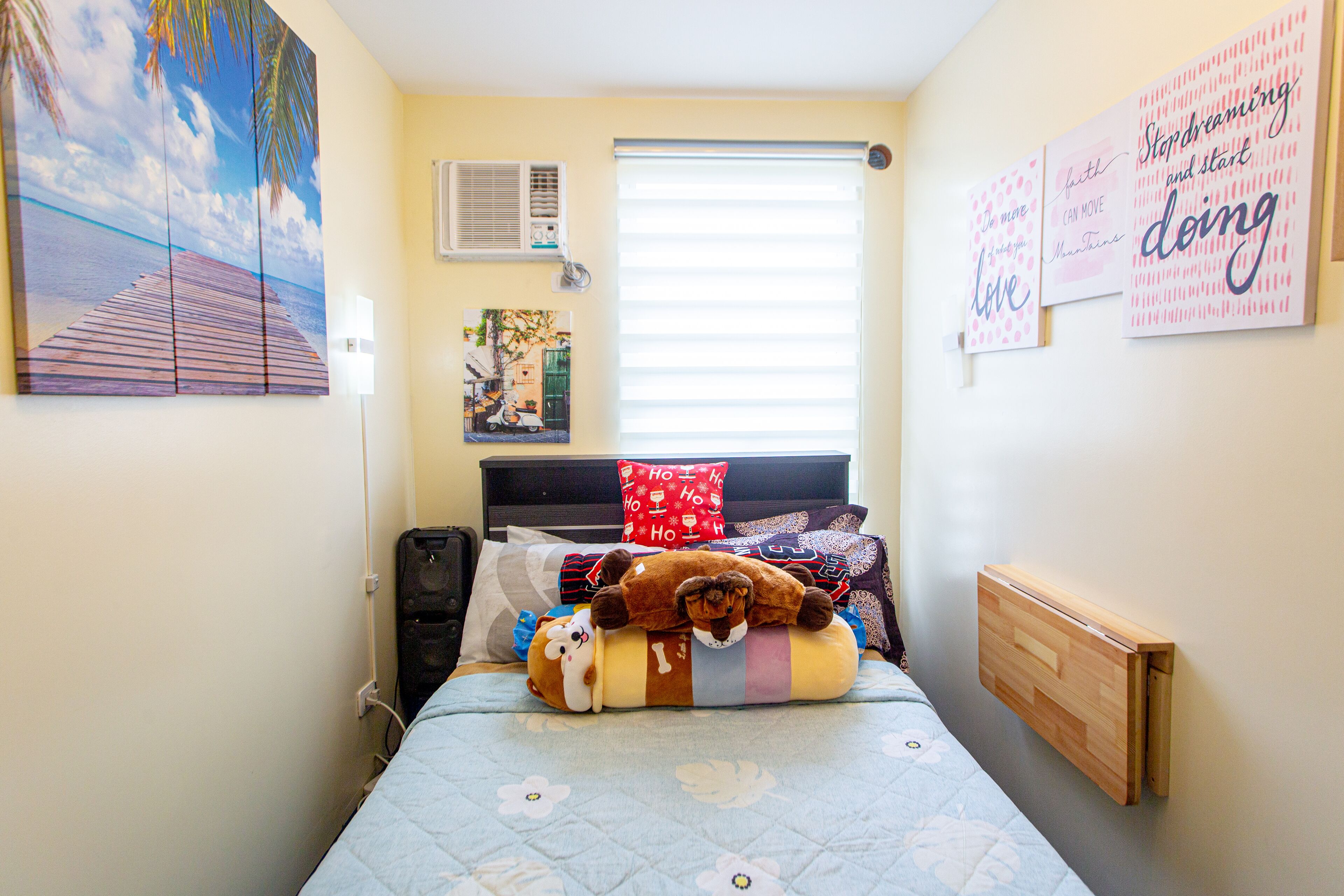 A bedroom with a bed, pillows, a stuffed toy, a wall-mounted air conditioner, a window with blinds, and motivational posters.
