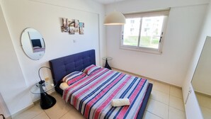 A bedroom with a bed, a round mirror, a bedside table, a lamp, and a window with a view of buildings. - Seaside Heaven Apartment (Larnaca)