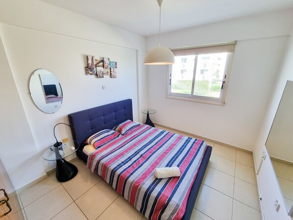 A bedroom with a bed, a round mirror, a bedside table, a lamp, and a window with a view of buildings.