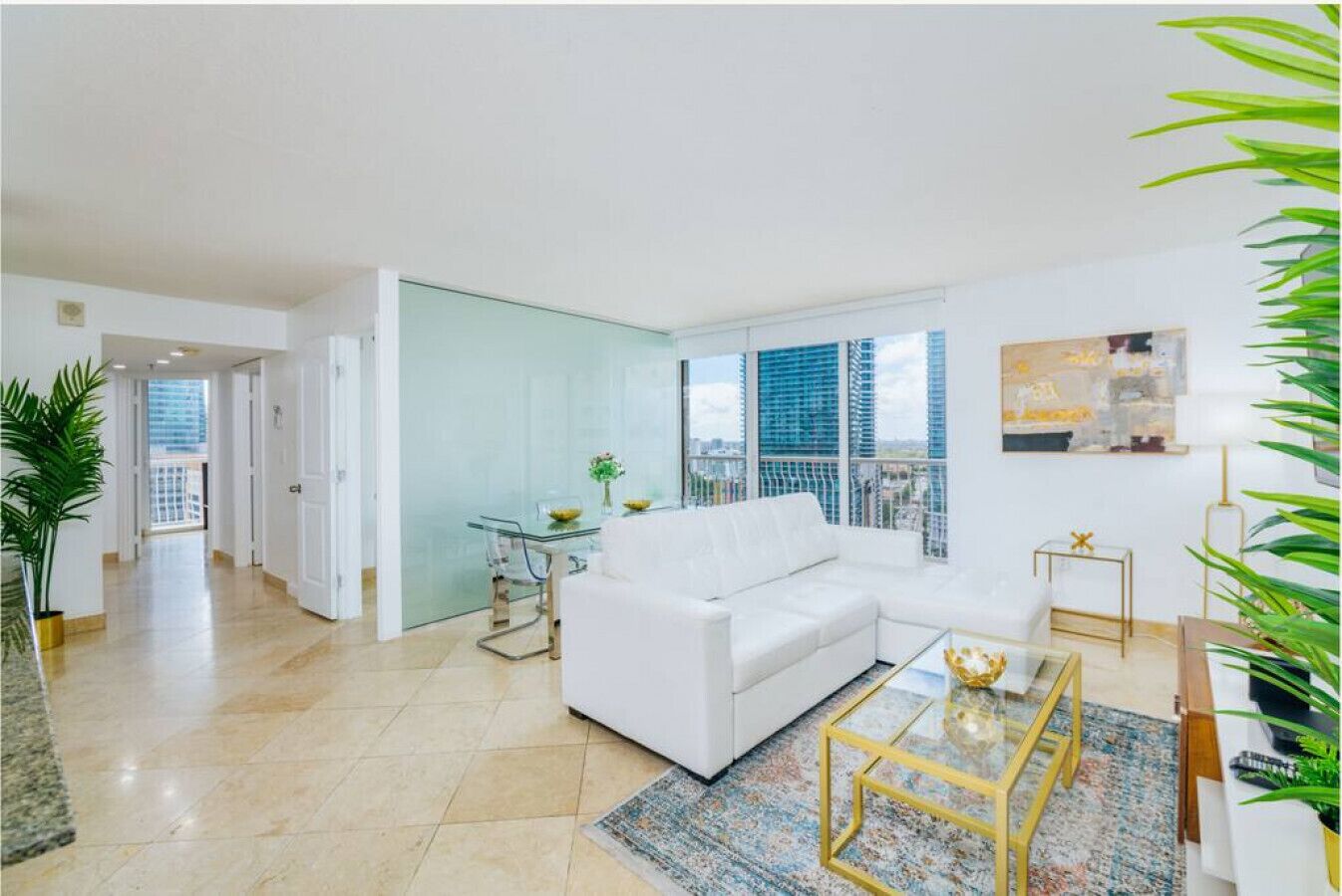 A modern living room with a white sofa, glass coffee table, and a view of the city through large windows.