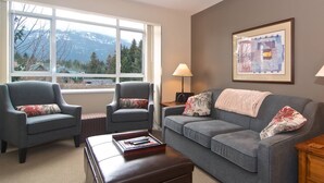 Sala de estar con dos sillones, un sofá, una mesa de centro y una lámpara. Se ve una ventana con vistas a las montañas y un cuadro enmarcado en la pared.