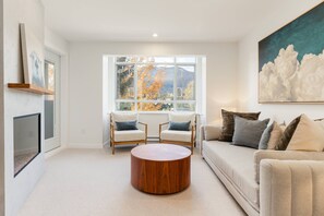 Ein Wohnzimmer mit großem Fenster, zwei Sesseln, einem runden Couchtisch und einer Couch mit vielen Kissen.