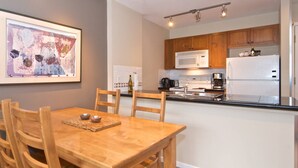 Un comedor con mesa y sillas de madera, una cocina con armarios de madera y electrodomésticos blancos, y un cuadro abstracto enmarcado en la pared.