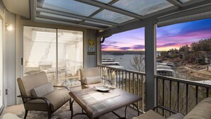A balcony with a glass enclosure, a table, chairs, and a view of a sunset over a body of water.
