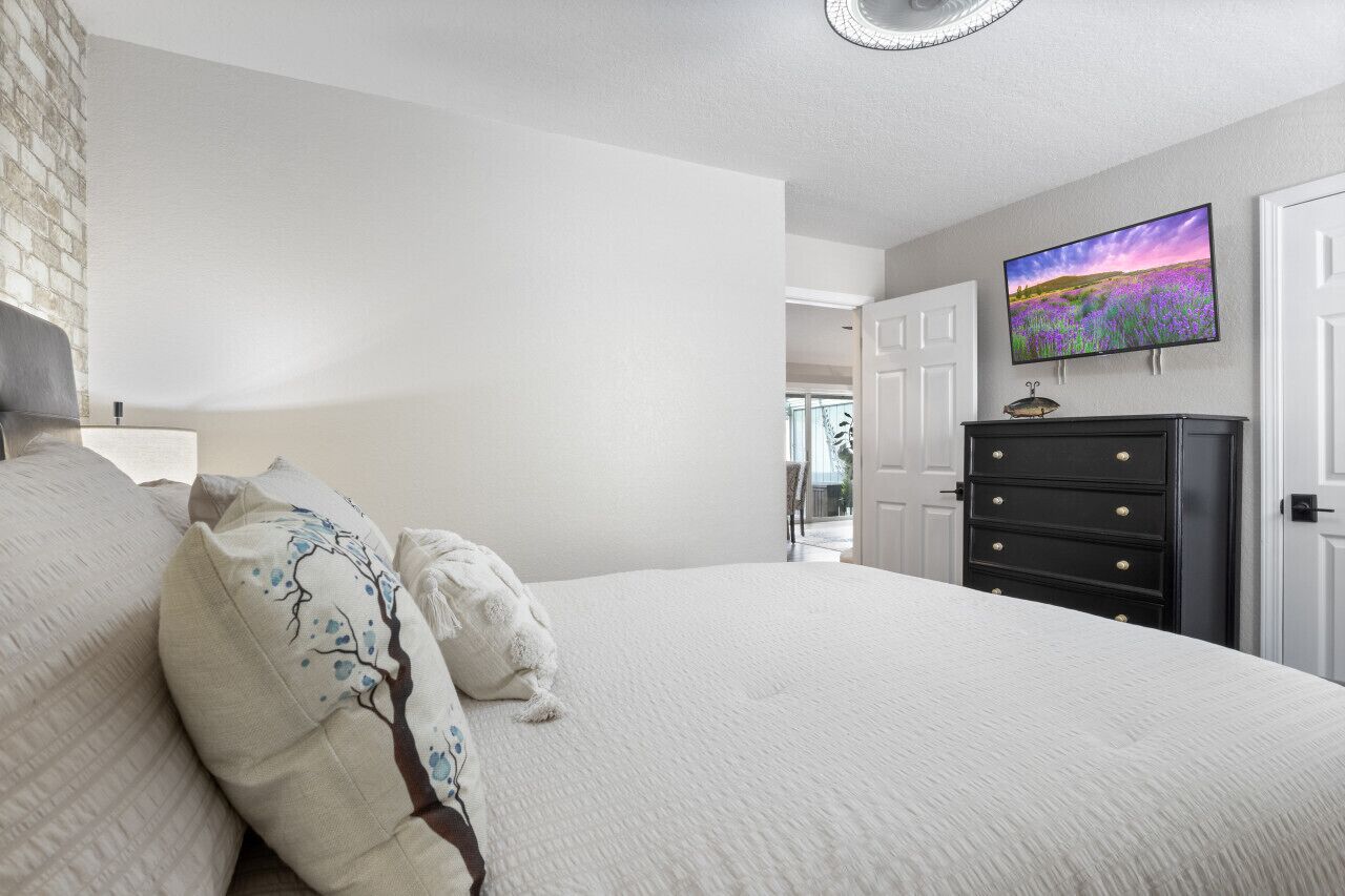 A bedroom with a large bed, a dresser, a TV mounted on the wall, and a door leading to another room.