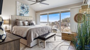 A bedroom with a large bed, a ceiling fan, a view of the outdoors, and a large window.