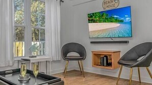 A modern living room with a large TV displaying a beach scene, two grey chairs, a wooden cabinet, and a glass of champagne on a tray. - 7BR Family Oasis | Heated Pool + Walk to Beach (North Myrtle Beach)