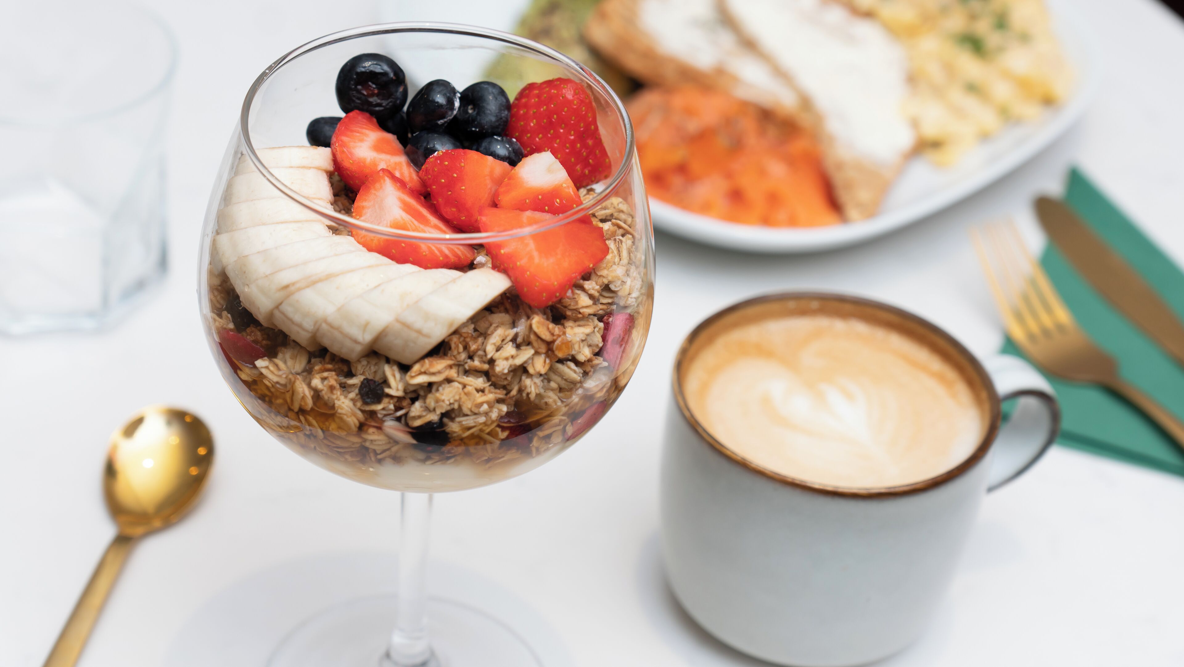 A dessert in a glass, a plate of food, a cup of coffee, and a spoon on a table.