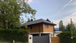 Ein modernes, zweistöckiges Haus mit grauer Garagentür, umgeben von viel Grün und blauem Himmel.