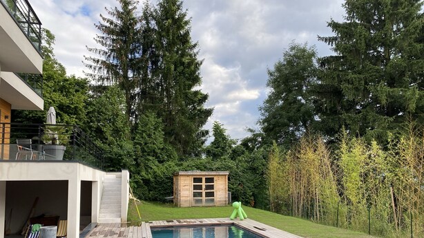 Ein Schwimmbecken mit Holzliegen, einer kleinen Holzhütte und einem üppigen Grün von Bäumen im Hintergrund.