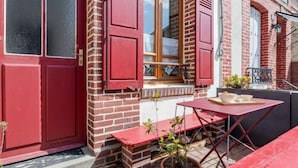 Un bâtiment en briques rouges, avec une porte rouge et des fenêtres fermées par des volets. Il y a un petit espace extérieur aménagé avec une table et des chaises, ainsi qu'un pot de fleurs.
