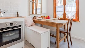 Une cuisine moderne avec des meubles blancs, un four encastré, une table à manger en bois avec des chaises, et une grande fenêtre donnant sur des immeubles.