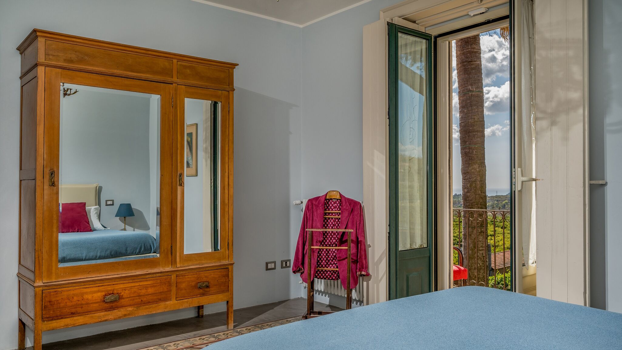 A bedroom with a large wooden wardrobe, a bed with blue bedding, and a view of a balcony with a palm tree.