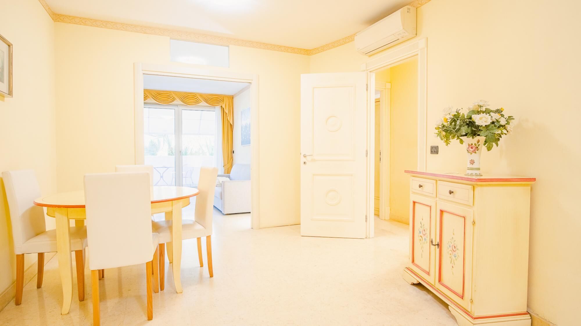 Uma área de refeições com uma mesa redonda e cadeiras, um armário branco com motivos florais e uma porta que dá para outro cômodo.