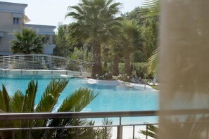 A swimming pool with a bridge, surrounded by palm trees and a building in the background. - 121 Relais du Lac Village - Italian Homing (Desenzano del Garda)