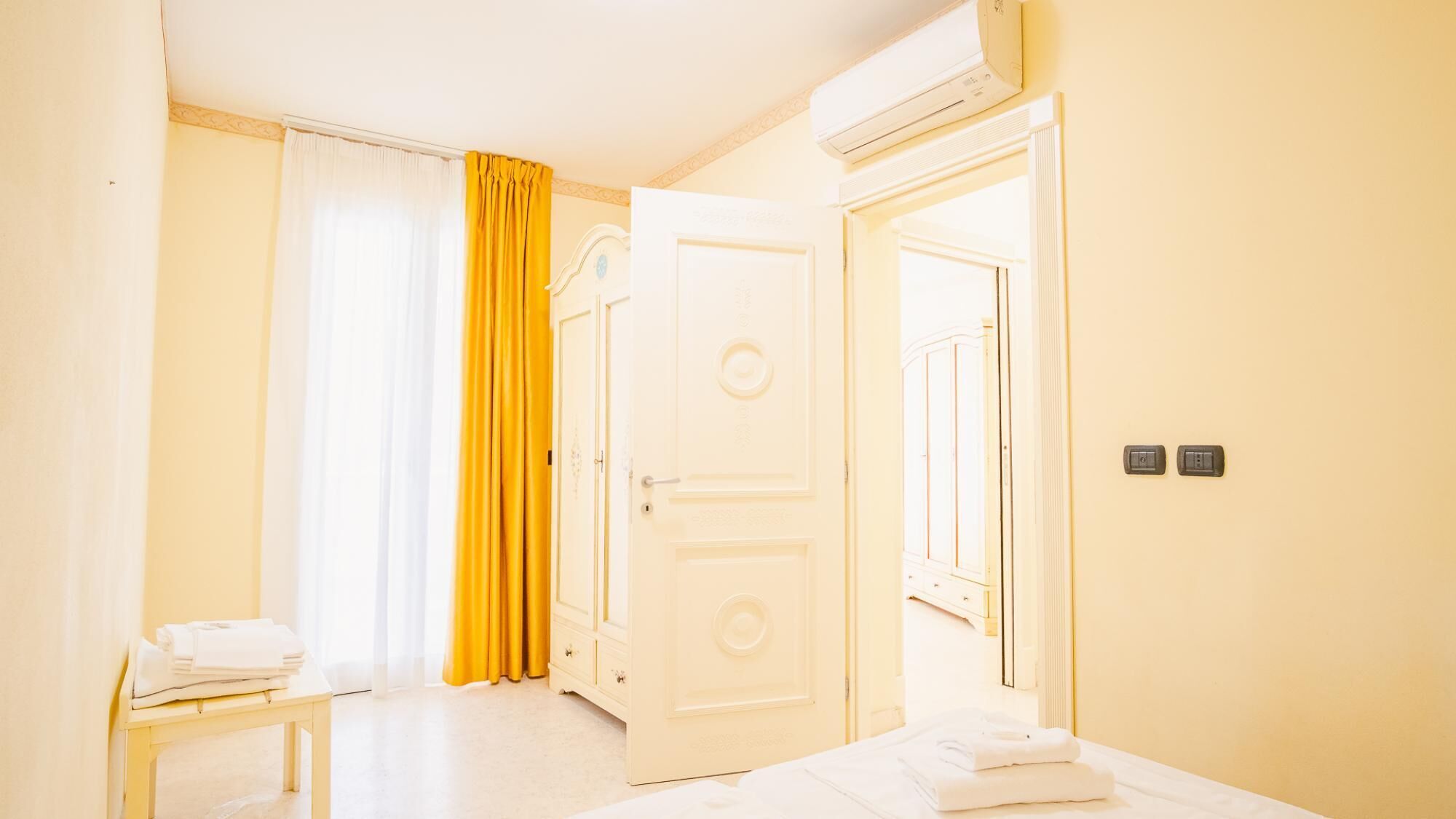 A hotel room with a bed, a bedside table with towels, a door, and an air conditioning unit.