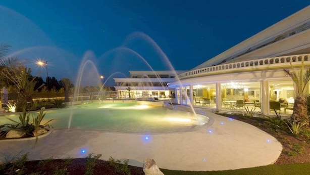 Un área de piscina exterior bien iluminada, con fuentes de agua, una zona de estar cubierta y un edificio con grandes ventanales.