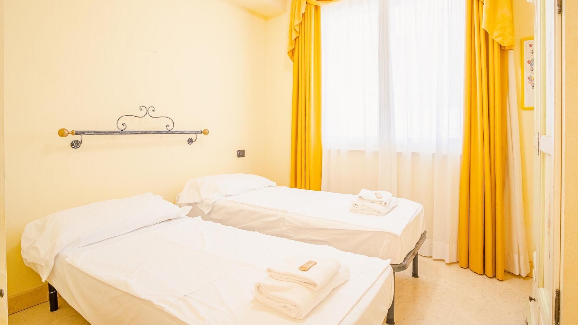 Una camera d'albergo con due letti singoli, una testiera in metallo e tende gialle.