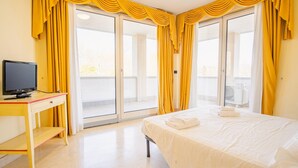 A hotel room with a bed, a TV, a desk, and a balcony with yellow curtains. - 104 Relais du Lac Village - Italian Homing (Desenzano del Garda)