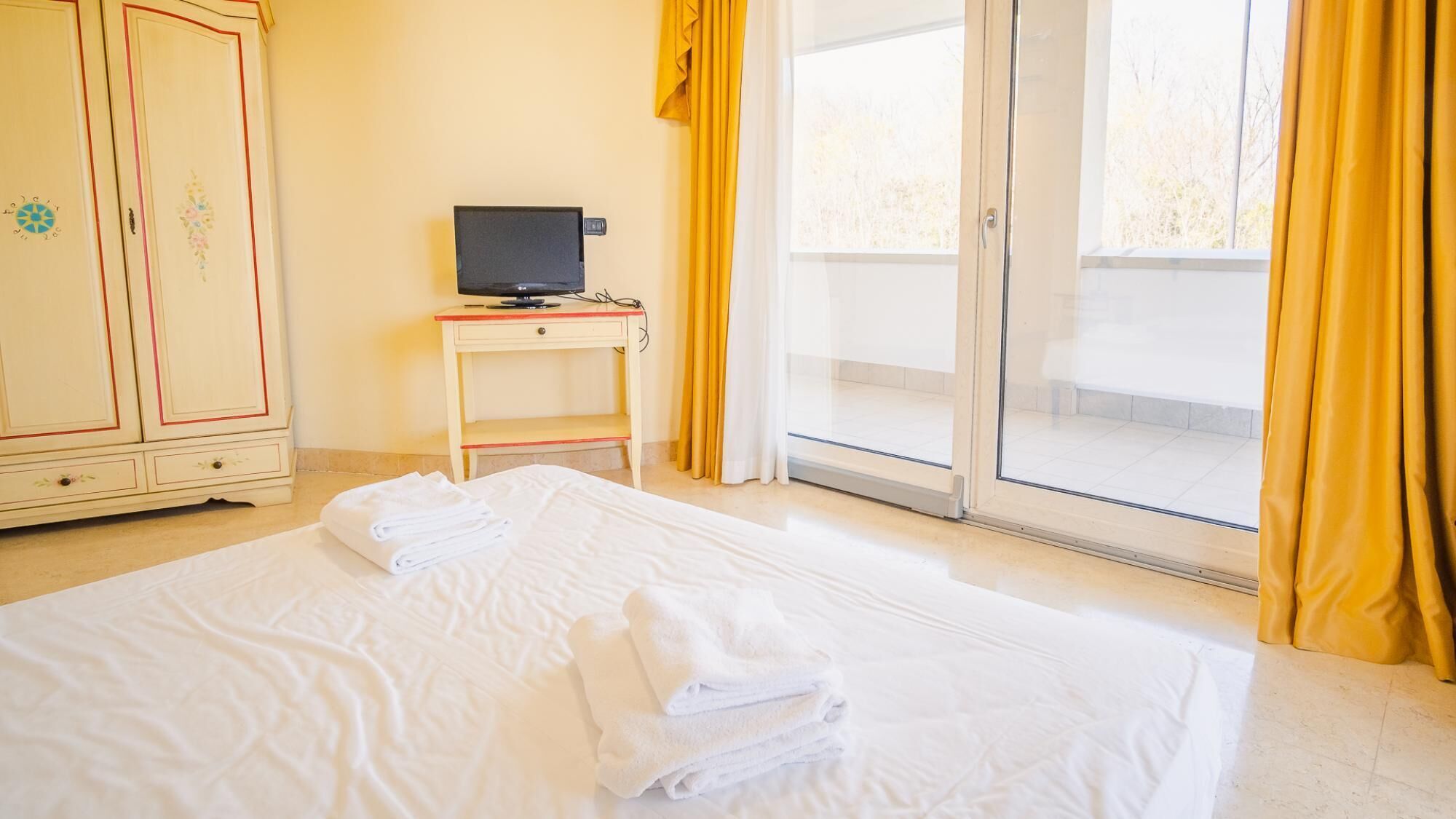 A hotel room with a bed, a TV on a small table, and a view of the outside through a glass door.
