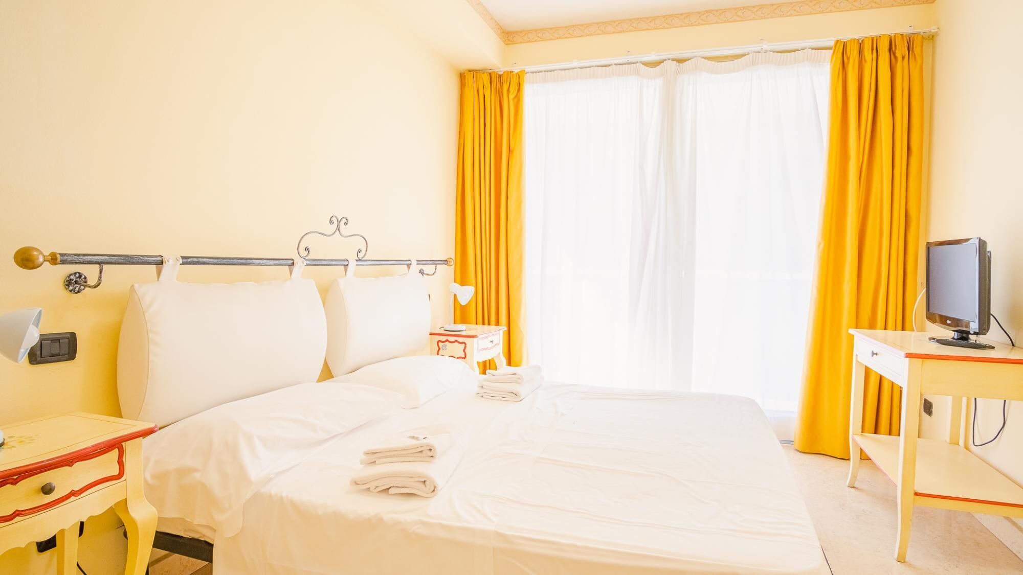 A hotel room with a bed, a desk with a computer, a lamp, and a window with yellow curtains.