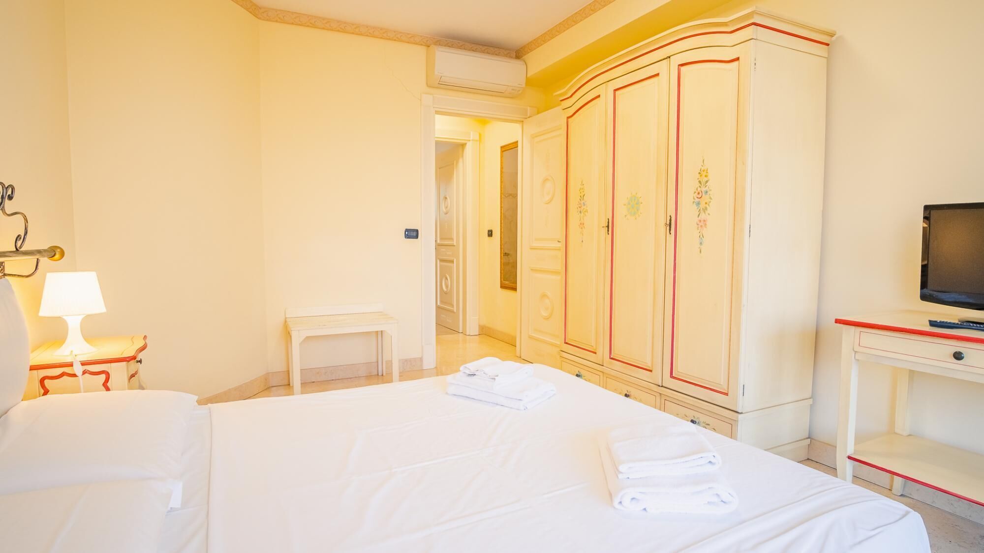 A hotel room with a large bed, a wardrobe, a television, and a small table with a lamp.