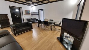 A modern living room with a black leather sofa, a glass dining table with chairs, a flat-screen TV, and a wooden floor.