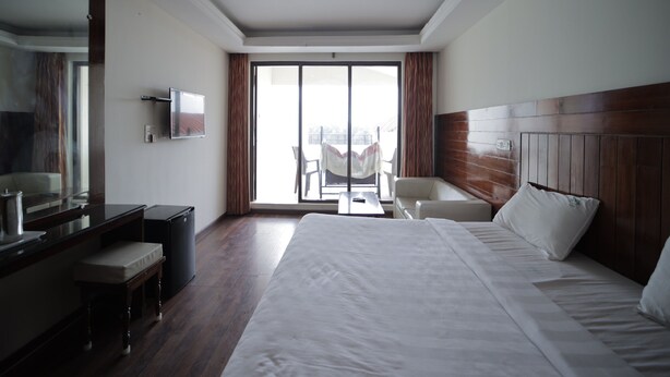 A hotel room with a large bed, a desk with a chair, a television, and a sliding glass door to a balcony. - Cool Stay Home (Kodaikanal)