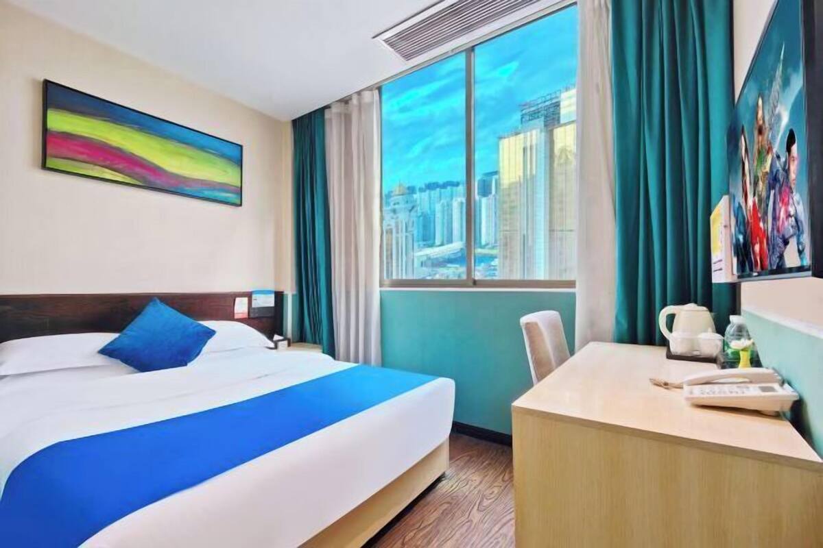 Une chambre d’hôtel avec un grand lit, un bureau, une vue sur la ville et un tableau coloré accroché au mur.