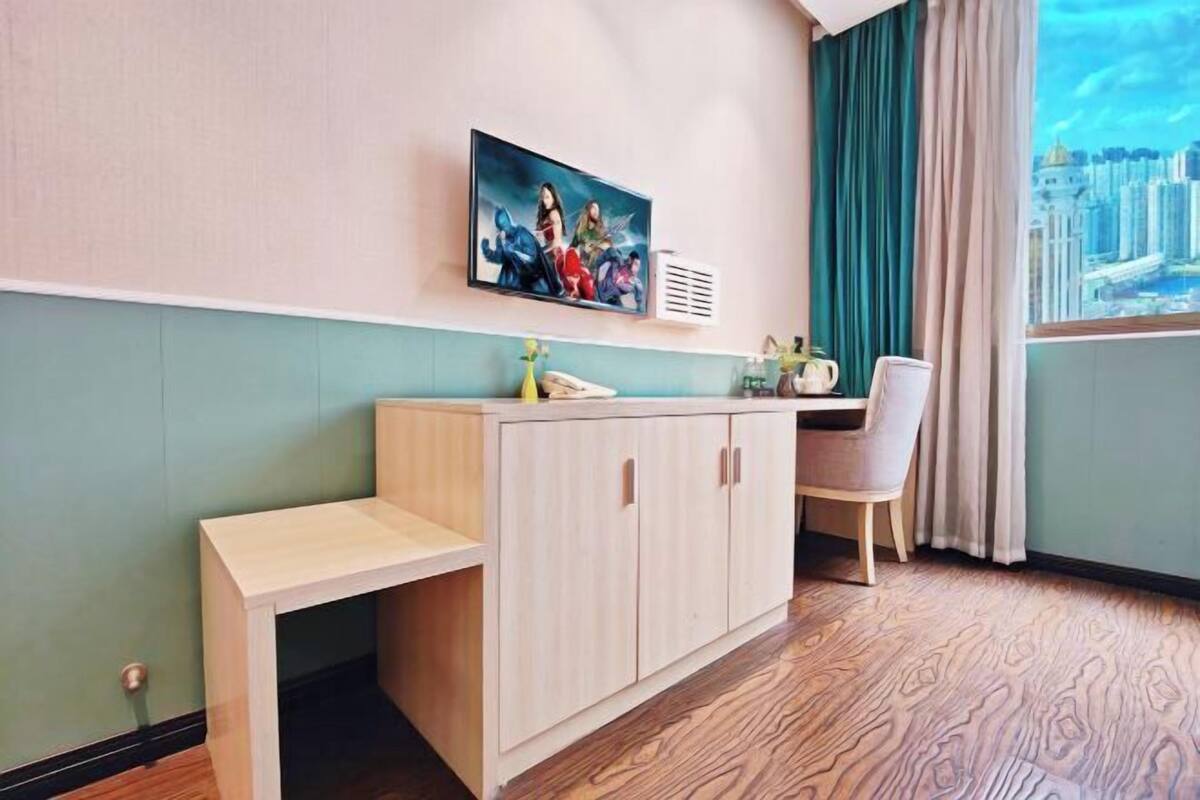 Ein Zimmer mit einem Holztisch, einem Stuhl, einem an der Wand befestigten Fernseher und einem Fenster mit Blick auf eine Stadtlandschaft.