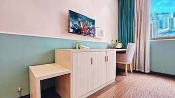 Une pièce avec un bureau en bois, une chaise, un téléviseur fixé au mur et une fenêtre donnant sur un paysage urbain.