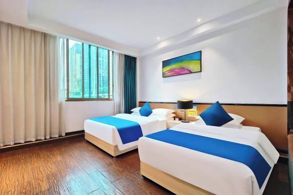 Une chambre d’hôtel avec deux lits, une grande fenêtre avec des rideaux, un tableau au mur et un plancher en bois.
