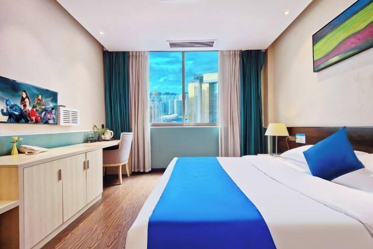 Une chambre d’hôtel avec un grand lit, un bureau et une vue sur le paysage urbain, visible à travers une grande fenêtre.